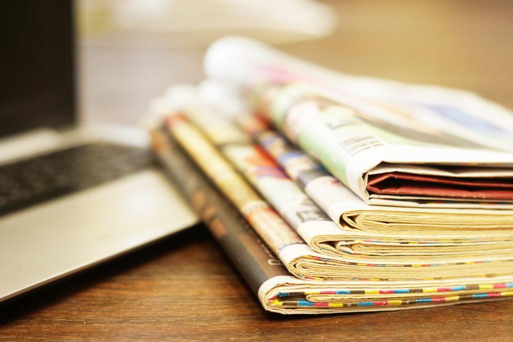 Das Foto zeigt einen Zeitungsstapel neben einem aufgeklappten Rechner